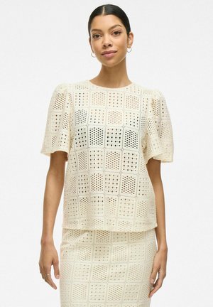 Top color crema con mangas cortas abullonadas, que presenta un patrón de rejilla con cortes. Textura suave y ajuste relajado, combinada con una falda a juego.