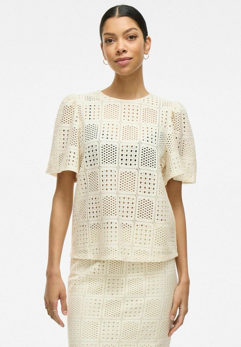 Top color crema con mangas cortas abullonadas, que presenta un patrón de rejilla con cortes. Textura suave y ajuste relajado, combinada con una falda a juego.