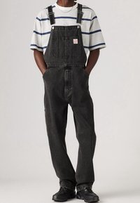 Svarta denimsoveraller med justerbara axelremmar, framlomme och sidolommer, med en randig vit och blå t-shirt och svarta sneakers.