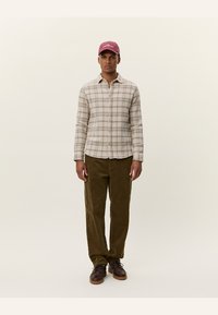 Plaid shirt in beige with green and brown accents, paired with olive corduroy pants and brown shoes. The model wears a burgundy cap.