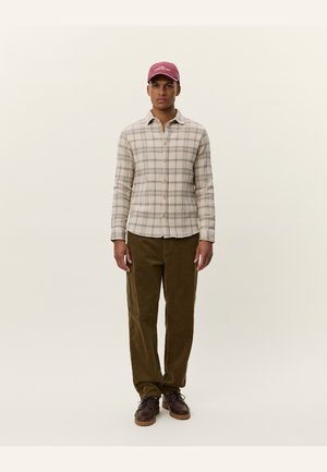 Plaid shirt in beige with green and brown accents, paired with olive corduroy pants and brown shoes. The model wears a burgundy cap.