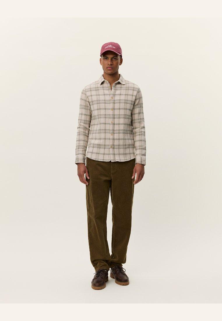 Plaid shirt in beige with green and brown accents, paired with olive corduroy pants and brown shoes. The model wears a burgundy cap.