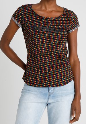 Frau trägt ein schwarzes kurzärmeliges Oberteil mit buntem, sich wiederholendem "DESIGUAL"-Text und schwarzem "Desigual"-Logo, kombiniert mit hellblauen Jeans.