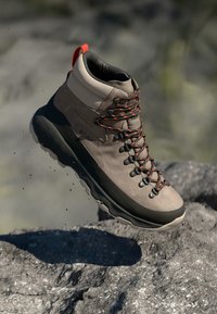 Grauer Wanderschuh mit strukturiertem Obermaterial, kontrastierenden roten Schnürsenkeln, gepolstertem Kragen und schwarzer Gummisohle. Über felsigem Gelände erhöht.