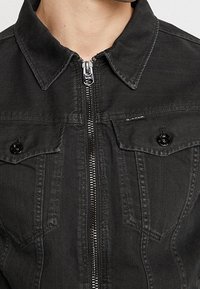 Giubbotto di jeans neri con zip frontale, due tasche sul petto con bottone e colletto a punta su un torso superiore di una persona.