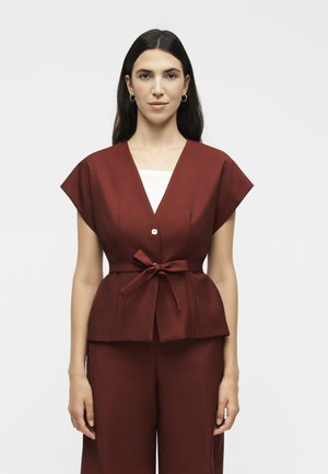 Γυναίκα με μακριά σκούρα μαλλιά που φοράει ένα μπουφάν με μανίκια μέχρι τον αγκώνα και ζώνη σε μπορντό χρώμα πάνω από λευκή μπλούζα και ταιριαστό φαρδύ παντελόνι μπορντό, σε όρθια στάση.