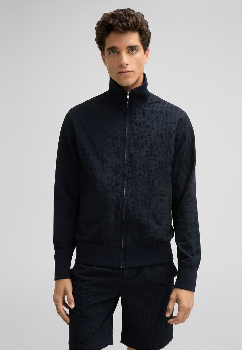 Marineblaue Zip-Jacke mit hohem Kragen, strukturiertem Stoff und gerippten Bündchen, kombiniert mit passenden Shorts. Minimalistisches Design ohne Muster.