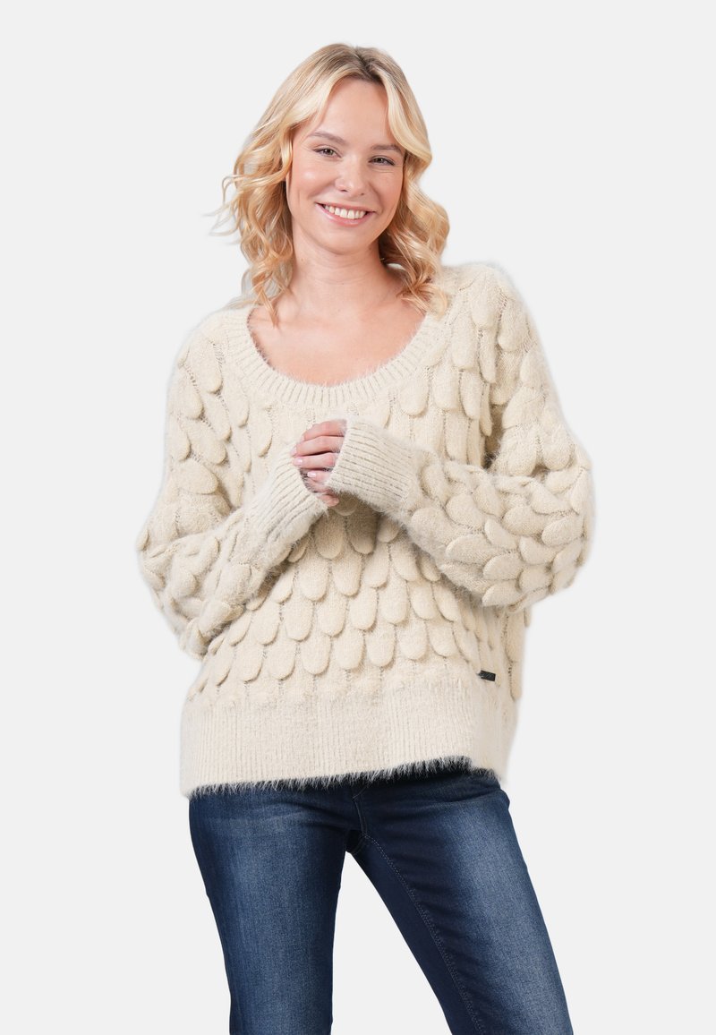 Cream-colored knitted sweater with a textured, bobble design, wide neckline, and ribbed cuffs, paired with dark blue denim jeans.