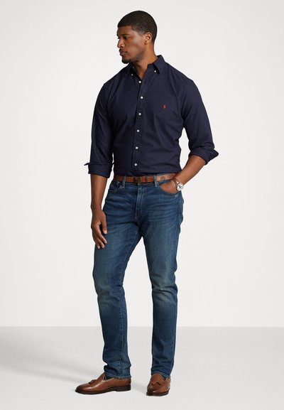 Chemise à manches longues bleu marine avec un petit logo, portée avec un jean foncé, une ceinture en cuir marron et des chaussures marron.