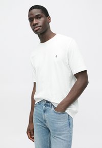 Balta īsroku T-krekls ar apaļu kakla izgriezumu un nelielu melnu simbolu kreisajā krūtīs, kombinēts ar gaiši ziliem džinsa džinsiem.