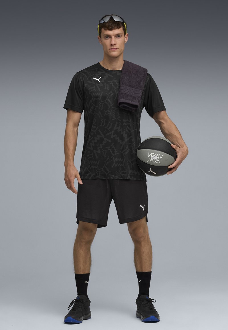 Homme athlétique vêtu de noir et portant des lunettes de soleil tient un ballon de basket dans une main et a une serviette posée sur l'épaule.