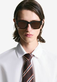 Giovane con capelli umidi di lunghezza media, che indossa occhiali da sole rettangolari scuri, una camicia bianca, una cravatta a righe marroni e un piccolo orecchino a cerchio.