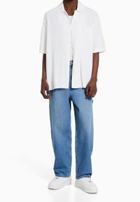 Osoba ubrana w białą koszulę z krótkim rękawem na guziki, białą koszulkę pod spód, jasnoniebieskie dżinsy, białe trampki oraz złoty pierścionek na lewej ręce.