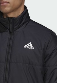 Giubbotto imbottito nero con colletto alto, chiusura a zip e logo Adidas bianco sul petto. Presenta una texture trapuntata e una finitura liscia.