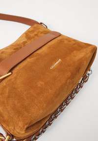 Geantă de umăr din suede maro cu o curea din piele, având un accesoriu din lanț metalic în partea de jos și detaliu cu logo embosat.