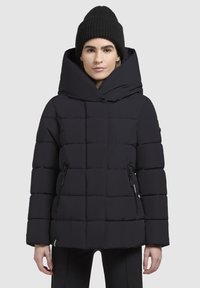 Schwarze Daunenjacke mit großer Kapuze, gestepptem Design, seitlichen Reißverschlüssen und einer glatten Textur. Begleitet von einer schwarzen Strickmütze.