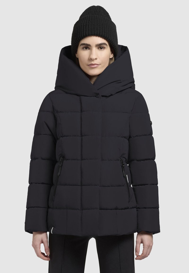 Schwarze Daunenjacke mit großer Kapuze, gestepptem Design, seitlichen Reißverschlüssen und einer glatten Textur. Begleitet von einer schwarzen Strickmütze.