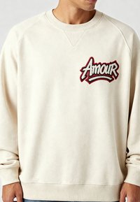 Sweat-shirt beige avec manches raglan, ornée d'une broderie "Amour" rouge et noire sur la poitrine. Tissu doux avec poignets et col côtelés.