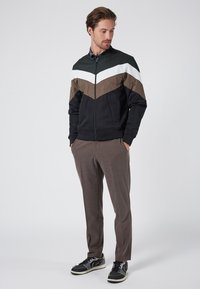 Giacca con zip nera, marrone, bianca e verde con design a chevron; abbinata a pantaloni marroni testurizzati e sneaker nere.