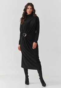 Czarna sukienka z długim rękawem, z przodem o skręconym designie, ozdobiona złotą klamrą w kształcie serca. Gładka, elastyczna tkanina, długość sięgająca kostek.