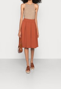 Nainen, jolla on yllään hihaton beige pusero, ruosteenvärinen pleatattu midi-hame, ruskeat kudotut alustan sandaalit ja joka kantaa ruskeaa teksturoitua käsilaukkua.