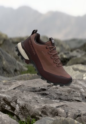 Brauner und dunkelweinroter Wanderschuh mit grober Sohle, Marke Scarpa, schwebt über felsigem Gelände mit verschwommenem Berg-Hintergrund.
