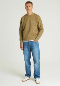 Felpa verde oliva con polsini a coste, scollatura rotonda e dettagli stampati. Abbinata a jeans azzurri chiari e sneakers bianche.
