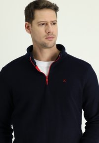 Marine-blauwe kwart-rits pullover gemaakt van zachte stof, met een rode rits en accent. Eenvoudig ontwerp met een klein logo op de borst.