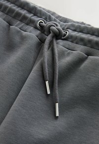 Tissu gris texturé avec une ceinture élastique et un cordon noué en nœud avec des embouts métalliques à chaque extrémité.