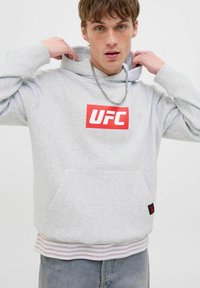 Junger Mann, der die Kapuze eines hellgrauen UFC-Hoodies anpasst, trägt eine silberne Kette, ein gestreiftes Hemd darunter und hellblaue Jeans.