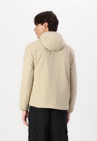 Beige lett jakke med hette, med lange ermer, glatt tekstur og en litt tettsittende design. Justerbare mansjetter synlige.