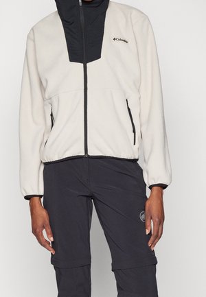Personne portant une veste polaire blanche Columbia avec une fermeture éclair noire et un pantalon d'extérieur noir avec des poches zippées.