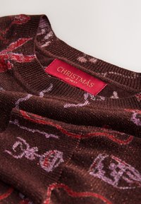 Pull marron tricoté avec une texture scintillante, arborant un motif sur le thème de Noël en tons rouges et violets. L'étiquette indique "NOËL chez NEXT."
