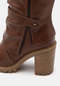 Brown faux leather ankle boot featuring a side zipper, adjustable strap, textured block heel, and ribbed outsole for traction.