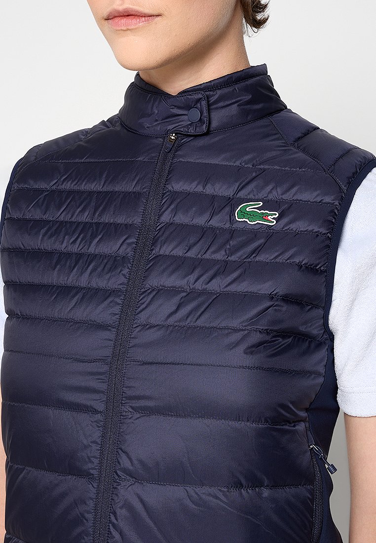Gilet matelassé bleu marine avec couture horizontale, col haut, fermeture éclair et logo brodé sur la poitrine gauche. Tissu lisse et léger.