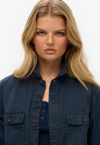 Chemise bleu marine à boutons avec un col montant, dotée de deux poches sur la poitrine et de boutons marron. Le tissu semble lisse et léger.