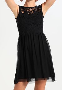Vestido negro con un cuerpo de encaje, mangas cortas y una falda de chiffon acampanada. El patrón de encaje presenta un diseño floral. Acentos ligeros y semi-transparentes.