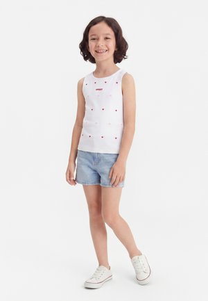 Lachend meisje met kort haar dat een witte mouwloze top draagt met kleine hartjes en de tekst "SWEET", lichte denimshorts en witte sneakers.