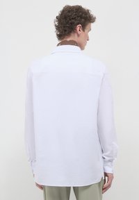 Camicia bianca a maniche lunghe con vestibilità comoda, caratterizzata da un yoke posteriore, orlo curvo e polsini con bottone. Tessuto liscio e leggero.