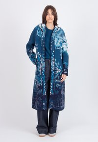 Blauwe gebreide jas met een capuchon, voorzien van een geometrisch patroon in verschillende tinten blauw. Gecombineerd met een blauwe top en donkere broek.