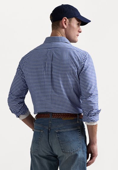 Blauw-wit geruite overhemden met lange mouwen, knoopsluiting bij de manchetten en een rugplooitje, gecombineerd met donkerblauwe jeans en een donkere pet.
