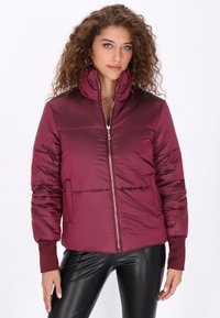 Bordó pufi dzseki magas gallérral, zipzárral és bordázott mandzsettákkal. Simogató, fényes textúrával és oldalsó zsebekkel rendelkezik.