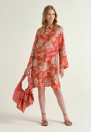 Donna che indossa un vestito lungo a maniche lunghe rosso con motivo floreale, che tiene una sciarpa rossa con fantasia e una borsa abbinata, in piedi davanti a uno sfondo semplice.