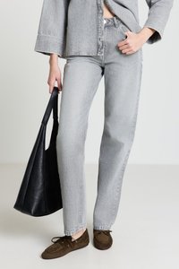 Graue Jeansjacke und passende Hose mit lockerem Schnitt, kombiniert mit dunkelbraunen Mokassins und einer schwarzen Tragetasche.