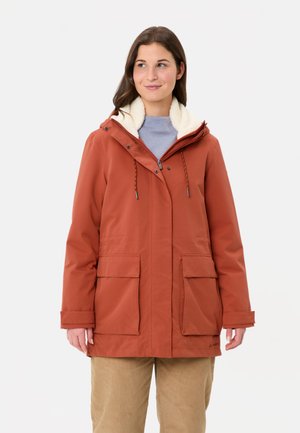 Vrouw in een roestkleurige winterjas met fleece voering, gedragen over een lichtblauw shirt en beige broek, staand tegen een witte achtergrond.