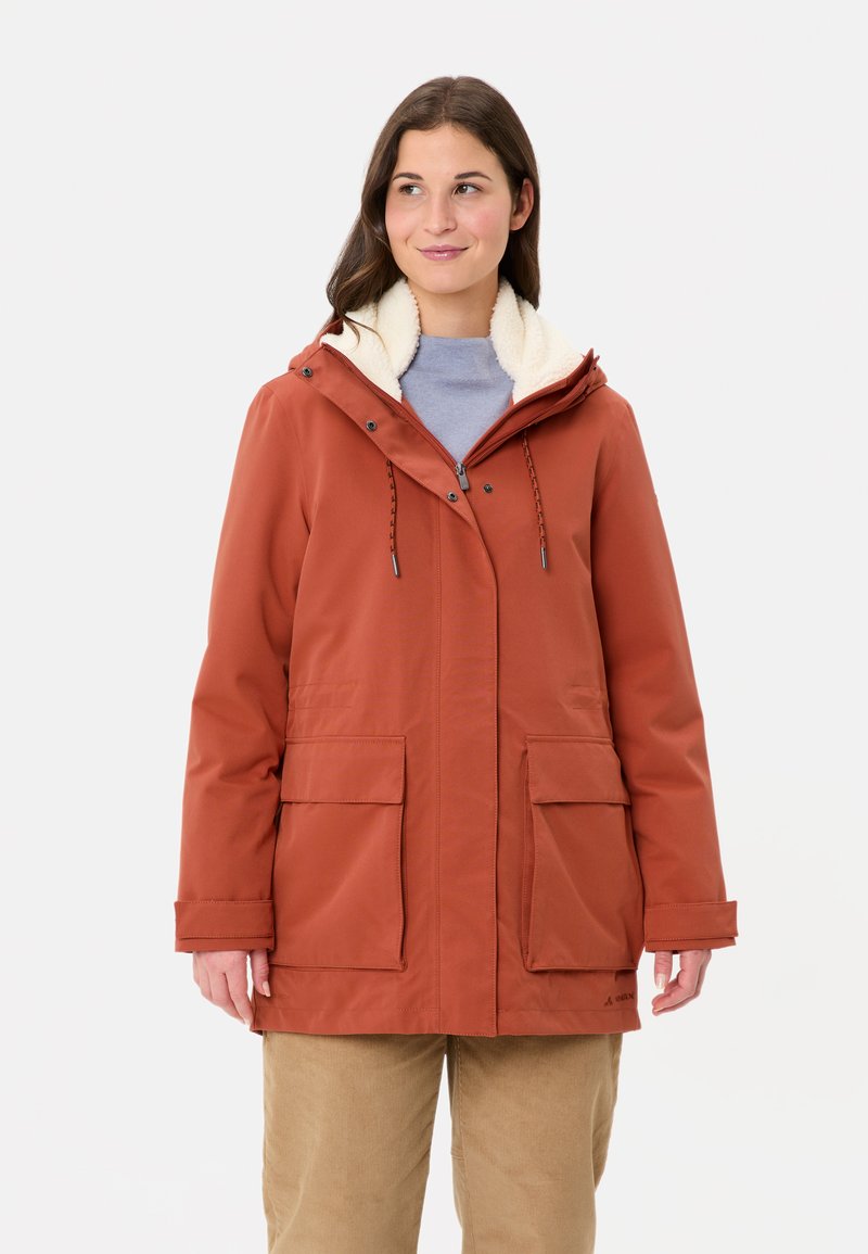 Donna che indossa una giacca invernale con cappuccio di colore ruggine, foderata in pile, sopra una maglietta azzurra chiara e pantaloni beige, in piedi contro uno sfondo bianco.