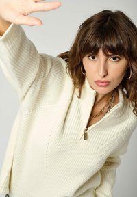 Pull en tricot couleur crème avec un col zippé mi-long, présentant une texture côtelée et un motif en chevrons subtil. Quincaillerie de fermeture éclair en ton doré.