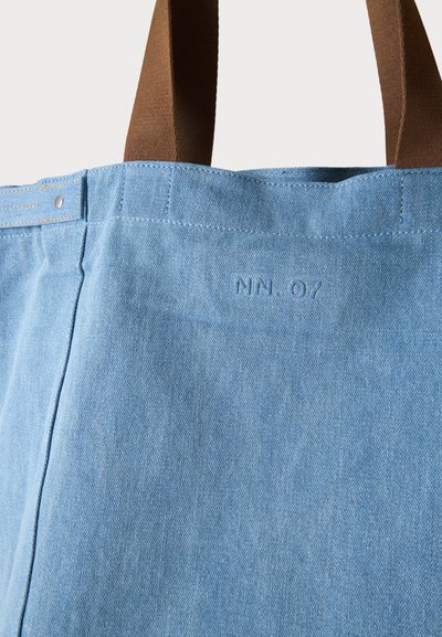 Sac fourre-tout en denim bleu clair avec poignées marron et lettrage discrètement cousu près du bord supérieur sur un fond uni.