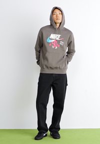 Grå hættetrøje med frontlomme, der har et farverigt grafisk design. Kombinationen er med sorte bukser og sorte sneakers.