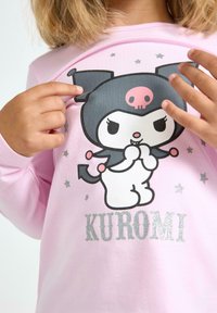 Kind in een roze shirt met een cartoonfiguur met zwarte oren, een roze schedel en de tekst "KUROMI" in zilveren letters.